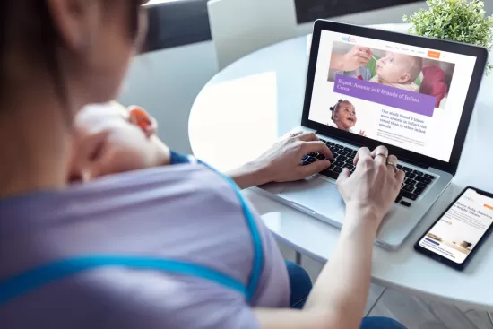 Woman with a baby in a carrier, browsing a website on her laptop